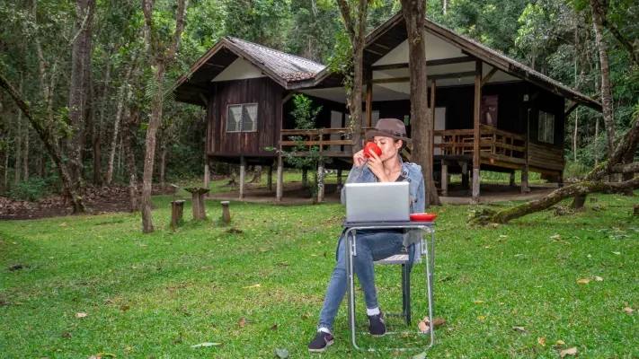 Žena pracuje na počítači a pije kávu před chatou v lese