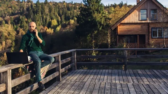 MUž s plnovousem sedí na teresa před chatou s notebookem a telefonuje