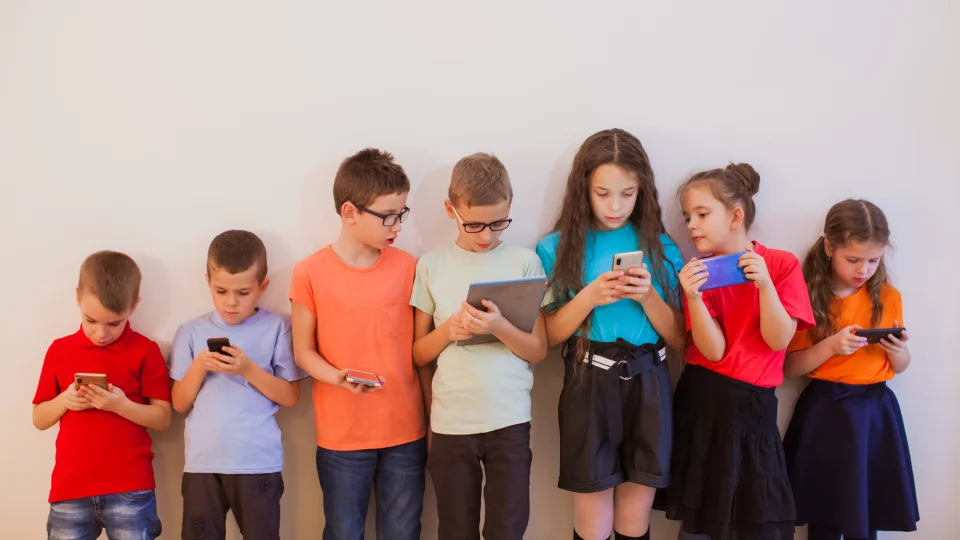 Skupinka dětí se opírá o bílou stěnu a sleduje obrazovky svých mobilů a tabletů