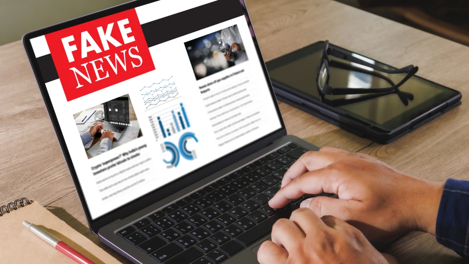 Detailní pohled na ruce na klávesnici notebooku. Na obrazovce je otevřená webová stránka s názvem Fake News.