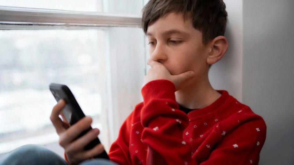 Chlapec sedí uvnitř u okna a sleduje obrazovku mobilního telefonu