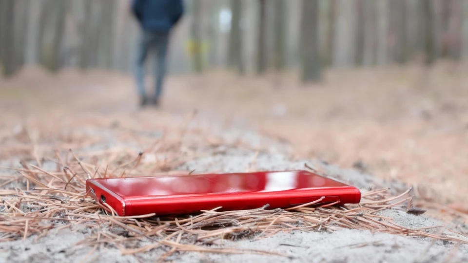 Detail na mobilní telefon pohozený na lesním podrostu, v pozadí silueta odcházející postavy