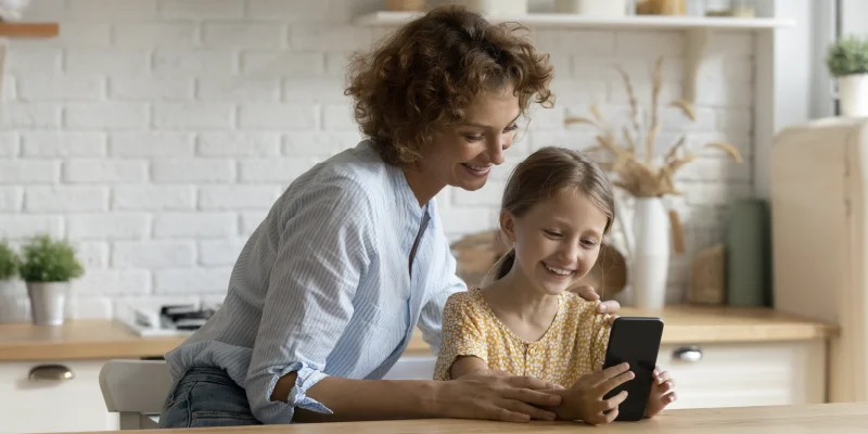 Matka a dcera společně sledují obrazovku mobilního telefonu v kuchyni